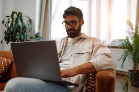 Photo for Handsome bearded man sitting on the couch by the window with a laptop, working online as a freelancer, shopping or chatting in the living room at home. - Royalty Free Image