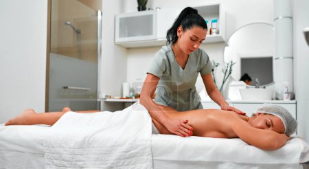 Téléchargez les photos : Gros plan de jeune femme détendue couchée face contre terre sur une table de massage et profitant d'un massage réparateur du corps effectué par un masseur professionnel dans un salon de spa ou un centre de bien-être - en image libre de droit