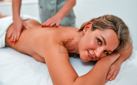Téléchargez les photos : Gros plan de jeune femme détendue couchée face contre terre sur une table de massage et profitant d'un massage réparateur du corps effectué par un masseur professionnel dans un salon de spa ou un centre de bien-être - en image libre de droit