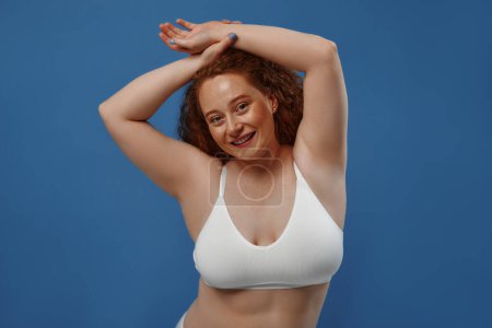 Photo for Young pretty redhead plus size or plump woman celebrating her natural body. Positive beautiful female model in white underwear against blue background. Lifestyle portrait with minimal makeup - Royalty Free Image
