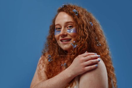 Photo for Beautiful redhead plus size woman with freckles and flowers in her hair using eye patches isolated on blue background. Body positive. Cosmetology and skin care. - Royalty Free Image