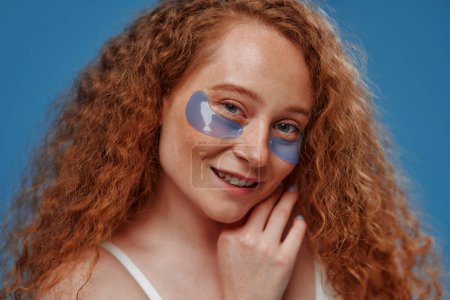 Photo for Beautiful redhead plus size woman with freckles and flowers in her hair using eye patches isolated on blue background. Body positive. Cosmetology and skin care. - Royalty Free Image