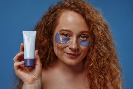 Photo for Beautiful redhead plus size woman with freckles using eye patches and holding tube of cream isolated on blue background. Body positive. Cosmetology and skin care. - Royalty Free Image