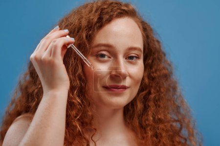 Photo for Freckled redhead plump plus size woman applying anti-wrinkle moisturizing serum on her face isolated on blue background. The concept of skin care, body positive and cosmetology. - Royalty Free Image