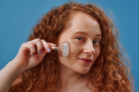 Photo for Freckled redhead plump plus size woman in white lingerie using face roller for facelift isolated on blue background. The concept of skin care, body positive and cosmetology. - Royalty Free Image
