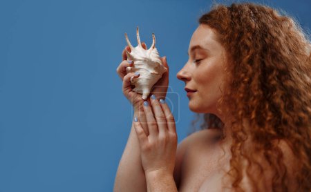 Photo for Freckled redhead plump plus size woman holding seashells isolated on blue background. The concept of face and body skin care, body positive and cosmetology. - Royalty Free Image