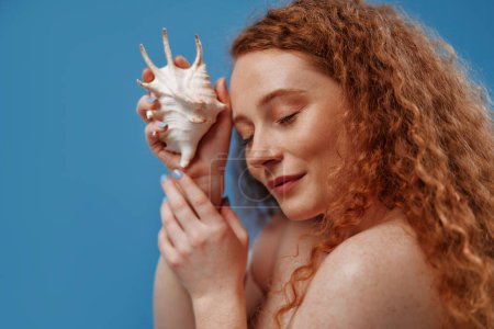 Photo for Freckled redhead plump plus size woman holding seashells isolated on blue background. The concept of face and body skin care, body positive and cosmetology. - Royalty Free Image
