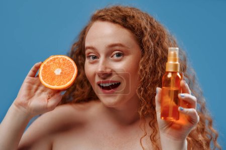 Photo for Freckled redhead plump plus size woman in white lingerie holding orange and jar of serum oil isolated on blue background. The concept of face and body skin care, body positive and cosmetology. - Royalty Free Image