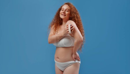 Photo for Freckled redhead plump plus size woman in white lingerie using anti-cellulite pilling massager brush isolated on blue background. The concept of body skin care, body positive and cosmetology. - Royalty Free Image