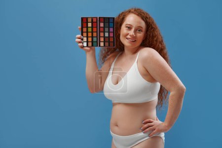 Photo for Beautiful plump plus size redhead woman with freckles holding eyeshadow palette isolated on blue background. - Royalty Free Image