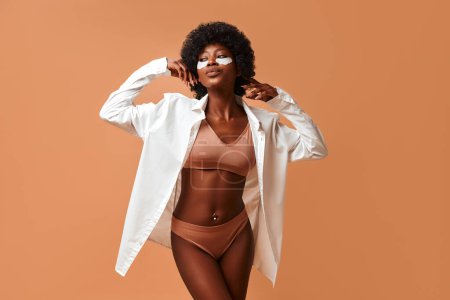 Téléchargez les photos : Belle femme afro-américaine utilisant des patchs hydratants en lingerie et chemise blanche posant en studio sur fond beige. Cosmétologie et soins de la peau spa. - en image libre de droit