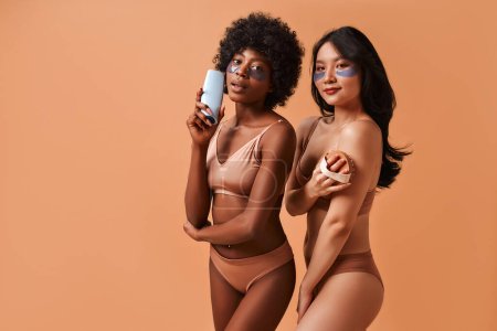 Photo for African-American and Asian women in lingerie using eye patches and holding a bottle of beauty product and anti-cellulite massage brush isolated on a beige background. Cosmetology and spa treatments. - Royalty Free Image