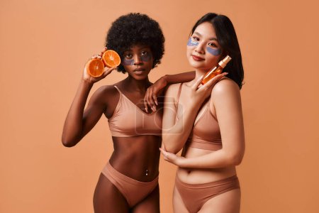 Foto de Mujeres afroamericanas y asiáticas en lencería usando parches para los ojos y sosteniendo fruta naranja y una botella de cabello o aceite corporal, el suero, aislado sobre un fondo beige. Cosmetología y tratamientos de spa. - Imagen libre de derechos