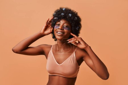 Téléchargez les photos : Belle jeune femme afro-américaine mince utilisant des patchs hydratants en lingerie en studio sur fond beige. Cosmétologie et spa concept de soins de la peau. - en image libre de droit