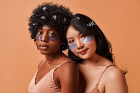 Téléchargez les photos : Jeunes femmes afro-américaines et asiatiques utilisant des bandeaux en lingerie posant sur fond beige. Cosmetologie et soins spa pour le visage et le corps. - en image libre de droit