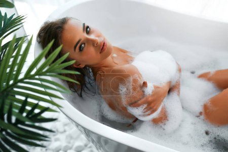 Photo for Beautiful passionate sexy sensual woman taking a relaxing bath with foam in a white bathroom by the window. Spa treatments for beauty and health with skin care - Royalty Free Image