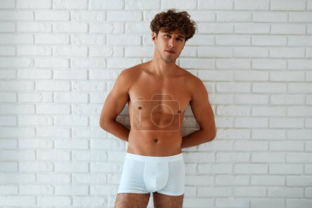 Photo for A handsome, muscular, sexy young man in white shorts with a naked torso standing against a white brick wall. - Royalty Free Image