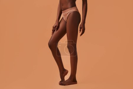 Téléchargez les photos : Portrait recadré des jambes d'une femme mince afro-américaine en lingerie beige isolée sur un fond beige. Concept de soins de la peau, de cosmétologie et de soins spa. - en image libre de droit