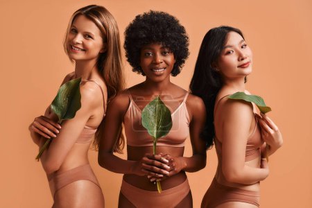 Téléchargez les photos : Femmes afro-américaines, asiatiques et caucasiennes en lingerie beige tenant une feuille verte posant sur fond beige. Le concept des soins de la peau, de la cosmétologie et des soins spa. - en image libre de droit