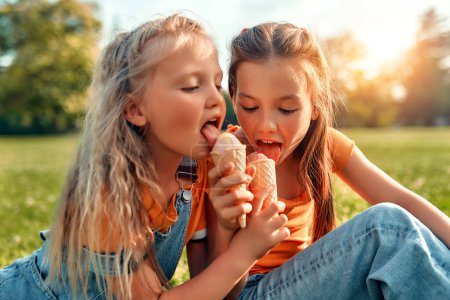 Foto de Dos hermanas comen helado mientras están sentadas en el césped en el parque en un día soleado y cálido. Dos niñas de niños jugando y comiendo dulces, sentadas en la hierba en el prado y descansando. - Imagen libre de derechos
