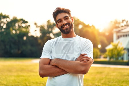 Photo for Handsome young bearded man in white t-shirt standing arms crossed in a meadow in a park on a sunny warm day, relaxing and enjoying nature on his day off. - Royalty Free Image