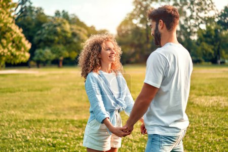Foto de Feliz joven hermosa pareja enamorada de pie en un prado en el parque tomados de la mano y disfrutando el uno del otro, mirando románticamente a los ojos en un cálido día soleado en su día libre. - Imagen libre de derechos