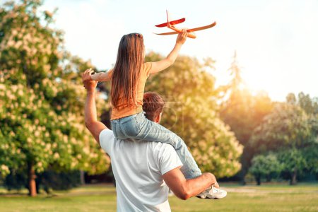 Foto de Feliz padre sosteniendo a su pequeña hija sobre sus hombros jugando con un avión en un prado en un parque en un cálido día soleado, divirtiéndose juntos en un día libre. - Imagen libre de derechos