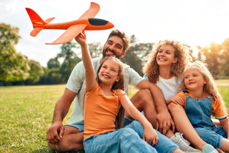 Foto de Feliz padre de familia joven, mamá y dos hijas niños jugando con un avión sentado en un prado en la hierba en el parque en un día cálido y soleado, divirtiéndose en un día libre. - Imagen libre de derechos