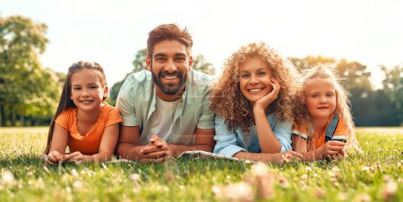 Photo for Happy young family dad, mom and two daughters lying on the grass in a meadow in the park on a warm sunny day, having fun together and relaxing on a day off. - Royalty Free Image