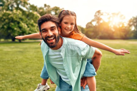 Photo for Happy young father playing with his little daughter, rolling her on his back, running and having fun in the meadow in the park on a warm sunny day on a weekend. - Royalty Free Image