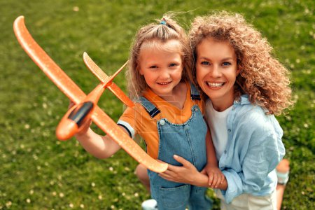Photo for Happy young mom playing with her beloved little daughter with an airplane in a meadow in the park, having fun together on a warm sunny weekend. - Royalty Free Image