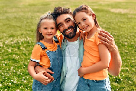 Photo for Happy young father hugging his two beloved cute daughters while sitting in a meadow in the park, playing and having fun on a warm sunny day off. - Royalty Free Image