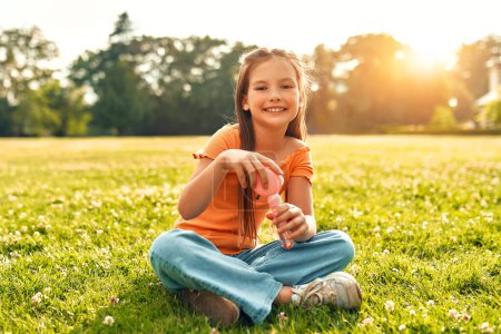 Photo for Cute beautiful girl blowing soap bubbles sitting on the grass in the meadow in the park, playing and having fun on a warm sunny weekend. - Royalty Free Image