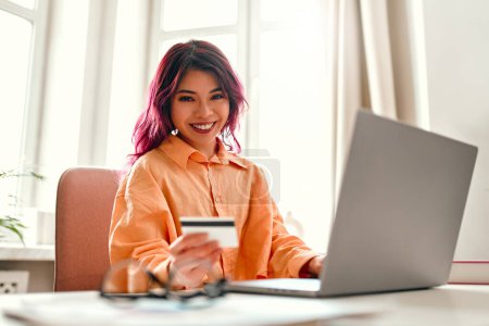 Foto de Chica joven asiática con hebras de color rosa brillante de pelo sentado en una mesa con un ordenador portátil en casa en la sala de estar, con una tarjeta de crédito. Compras en línea y banca. - Imagen libre de derechos
