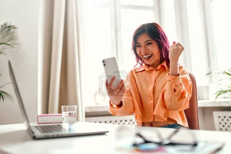 Photo for Asian young girl with bright pink strands of hair sitting at a table with a laptop at home in the living room, shocked looking at the smartphone and showing yes gesture. - Royalty Free Image