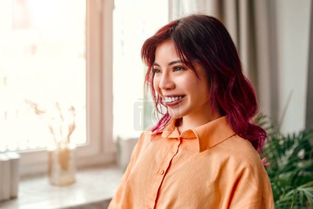 Photo for Close-up portrait of a young bright Asian woman with pink streaks of hair in a shirt standing by the window at home in the living room or office. - Royalty Free Image