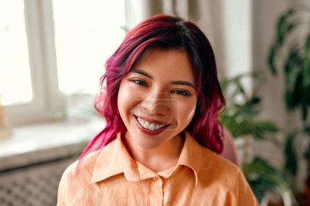 Téléchargez les photos : Portrait en gros plan d'une jeune femme asiatique brillante avec des rayures roses de cheveux dans une chemise debout près de la fenêtre à la maison dans le salon ou le bureau. - en image libre de droit