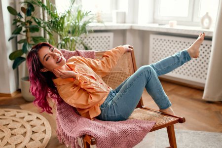 Photo for Portrait of a young bright Asian woman with pink strands of hair in a shirt sitting in a chair with her legs up and relaxing in the living room. - Royalty Free Image