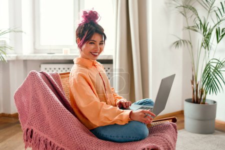 Photo for Portrait of a young bright Asian woman with pink streaks of hair sitting in a chair with a laptop and taking online training or working in the living room. - Royalty Free Image