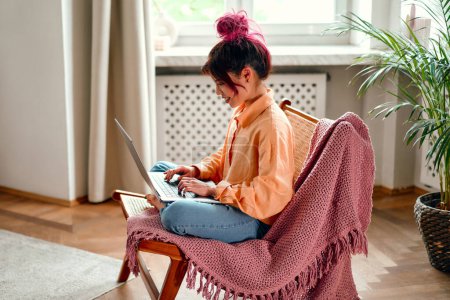 Téléchargez les photos : Jeune femme asiatique avec des rayures roses vives de cheveux assis dans un fauteuil confortable avec un ordinateur portable dans le salon à la maison tout en travaillant ou en suivant une formation en ligne. - en image libre de droit