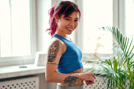 Photo for Young Asian woman with bright pink streaks of hair and tattoos in sportswear exercising at home in the living room. Sports, yoga and active lifestyle. - Royalty Free Image