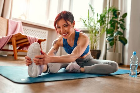 Foto de Mujer asiática joven con tatuajes en ropa deportiva ejercitándose en casa en la sala de estar en una alfombra de goma haciendo ejercicios de estiramiento. Deportes, yoga y estilo de vida activo. - Imagen libre de derechos
