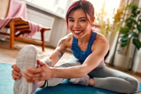 Foto de Mujer asiática joven con tatuajes en ropa deportiva ejercitándose en casa en la sala de estar en una alfombra de goma haciendo ejercicios de estiramiento. Deportes, yoga y estilo de vida activo. - Imagen libre de derechos