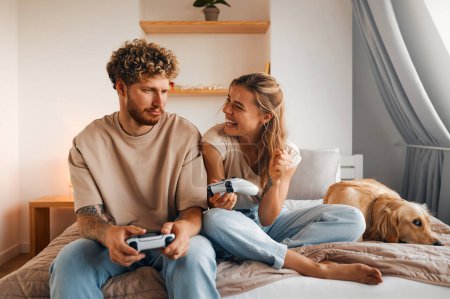 Téléchargez les photos : Jeune couple amoureux jouant à des jeux vidéo tout en étant assis sur le canapé dans la chambre à coucher, s'amuser et profiter l'un de l'autre le week-end. - en image libre de droit