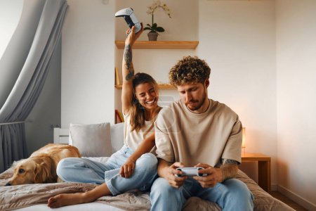 Photo for Young couple in love playing video games while sitting on the sofa in the bedroom, having fun and enjoying each other on their weekends. - Royalty Free Image