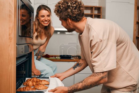 Foto de Pareja joven enamorada horneando croissants en la cocina juntos, preparándose para el desayuno, hablando y pasando tiempo juntos románticamente. - Imagen libre de derechos