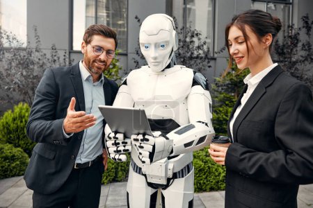 Photo for Young man and woman business partners in suits with a tablet standing together with a robot and discussing work against the background of a building. The future with artificial intelligence. - Royalty Free Image
