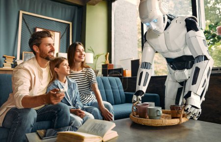 Foto de Una familia con una hija sentada en el sofá de la sala de estar usando una tableta y un robot que les preparaba té y café en una bandeja. Vivir juntos entre humanos e inteligencia artificial. - Imagen libre de derechos