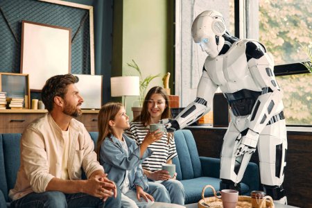 Photo for A young family with a daughter sitting on the sofa in the living room and a robot housekeeper who serves them tea and coffee. Living together between humans and artificial intelligence. - Royalty Free Image