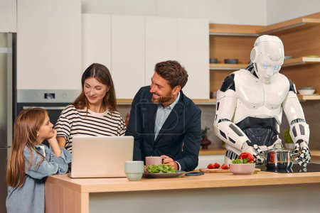 Foto de Robot ama de llaves preparando la comida en la cocina, mientras que la familia con la hija usando el ordenador portátil. Vivir juntos entre humanos e inteligencia artificial. - Imagen libre de derechos